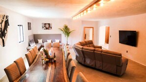Uma sala de estar ampla com uma grande mesa de jantar de madeira, um sofá modular e uma TV de tela plana fixada na parede.