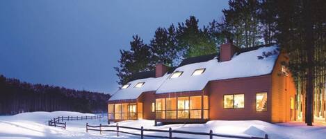 Une maison moderne en bois, à plusieurs étages, dotée de grandes fenêtres et d’un toit enneigé, entourée d’une clôture en bois et d’arbres.
