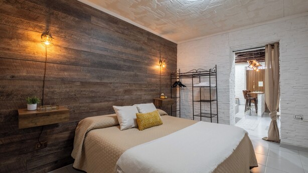 A modern bedroom with a wooden wall, a bed with a white and beige bedspread, a hanging light fixture, and a small plant on a shelf. - Su Lizu, charm and relaxation in the historic center of Cagliari - with patio. (Cagliari)