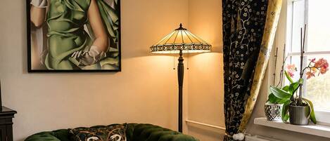 Um quarto com um sofá estofado verde, uma mesa redonda com toalha de mesa floral, um abajur, um quadro na parede e uma janela com cortinas.