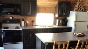 Une cuisine avec des meubles de rangement en bois foncé, un réfrigérateur blanc, un four à micro-ondes, une cuisinière et un évier. On y trouve deux tabourets de bar en bois et un panier de fruits sur le comptoir.