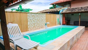 Ein rechteckiger Pool mit einer bepflasterten Kante, ein weißer Liegestuhl und ein Holzzaun.