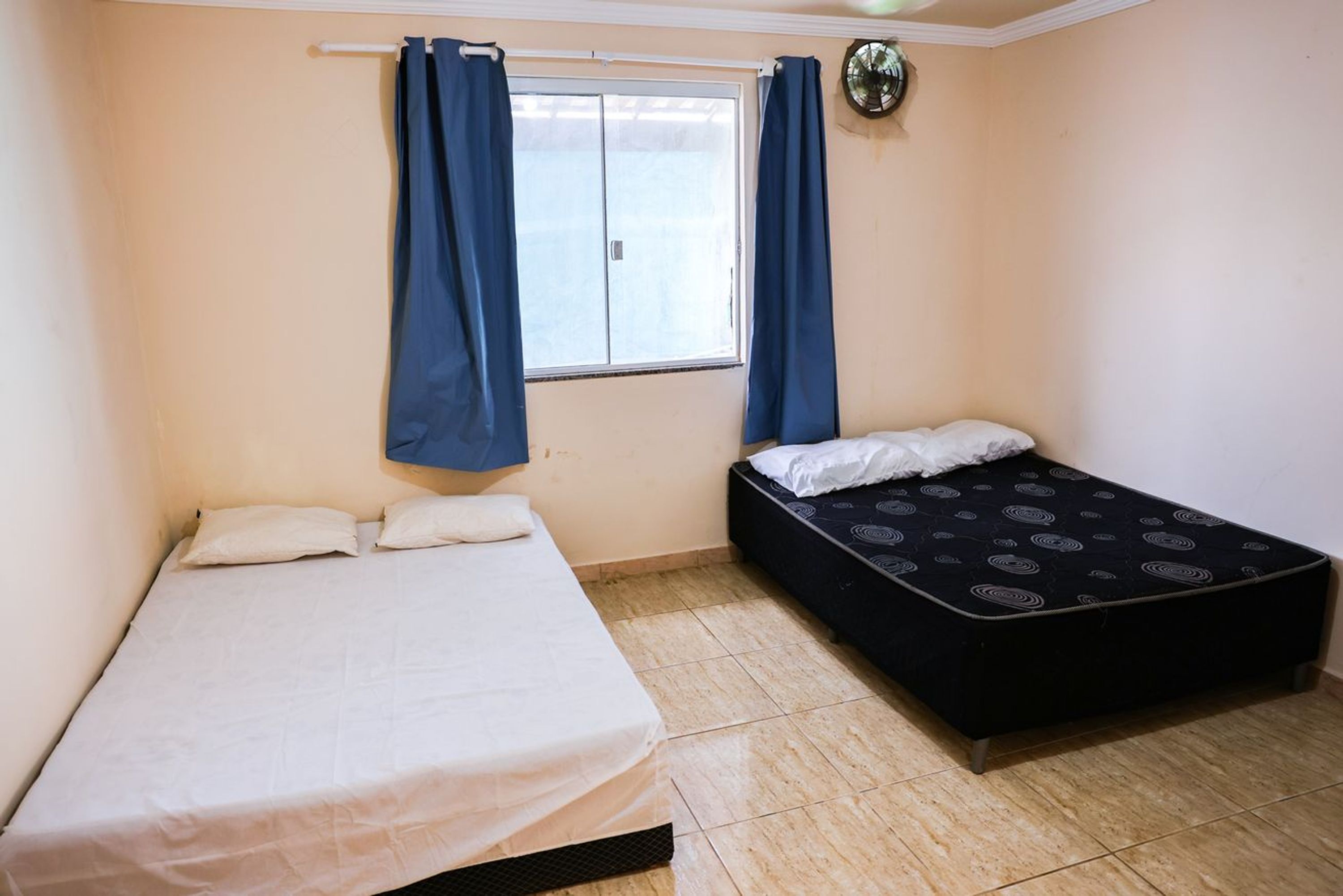 Une chambre avec deux lits, l’un avec un matelas blanc et l’autre avec un matelas noir à motifs. Il y a une fenêtre avec des rideaux bleus, un ventilateur de plafond et un sol carrelé.