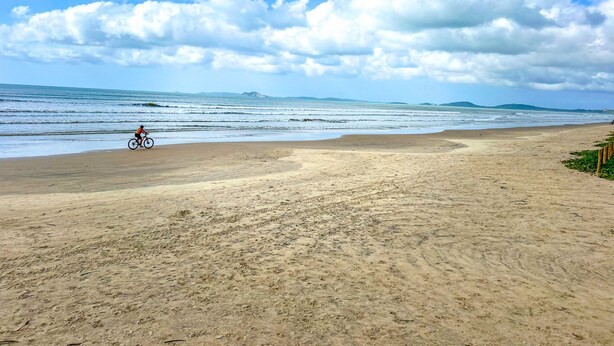 Ein Strand mit einem Radfahrer, sandigem Untergrund und einem klaren Himmel mit Wolken.