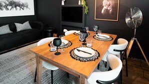 A dining area with a wooden table set for four, surrounded by white chairs. There are two pendant lights with a woven design above the table. The room has a black wall with a framed picture of a tiger and a black sofa with patterned cushions.