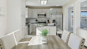 A modern kitchen with white cabinetry, stainless steel appliances, and a dining area with a wooden table and white chairs. - Hooked on Avocado -Stunning 5 Bedroom/ 3 Bath waterfront home on canal, resort style private pool & hot tub, 34’ dock, and quick open-water Gulf and Atlantic access. Ice Maker, Ping Pong table. Minutes to Sombrero Beach & Dining. (Marathon)