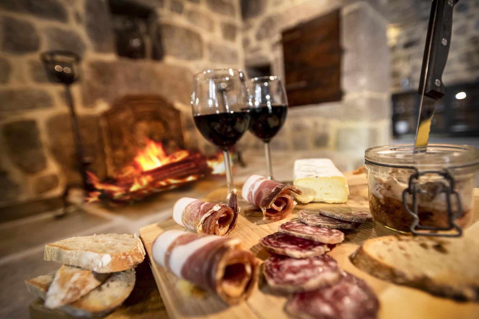 A charcuterie board with various meats, cheese, and bread, accompanied by glasses of red wine, set against a stone fireplace.
