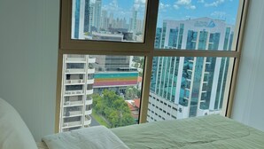 Ein Schlafzimmer mit großem Fenster und Blick auf die Stadt, ein Bett mit weißer Bettwäsche und Hochhäuser im Blick.