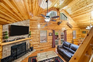 A log cabin interior with a fireplace, leather sofa, ceiling fan, and a television mounted on the wall. - Smoky Mtn Cabin w Hot Tub & Game Room! (Sevierville)