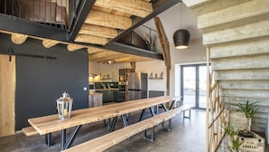 Un vaste espace à manger avec une longue table en bois et des bancs, une cuisine à l’arrière-plan et un plafond haut avec des poutres apparentes.