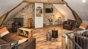 Une pièce mansardée chaleureuse avec des poutres apparentes, des murs en pierre et une cheminée. On y trouve des canapés en cuir et une table basse en bois.