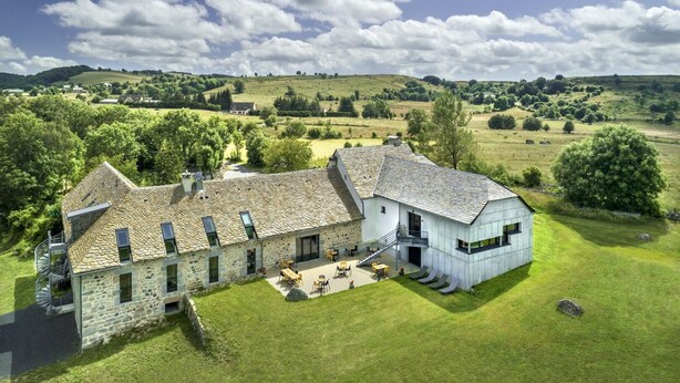 Un bâtiment en pierre doté d’une extension moderne, entouré d’une pelouse bien entretenue et d’arbres, situé dans un paysage rural.