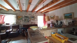 A rustic interior with exposed wooden beams, a stone wall, a dining area with a wooden table, and a kitchen with a sink and stove. - L'Oustal de la Fillo (CHAUCHAILLES)