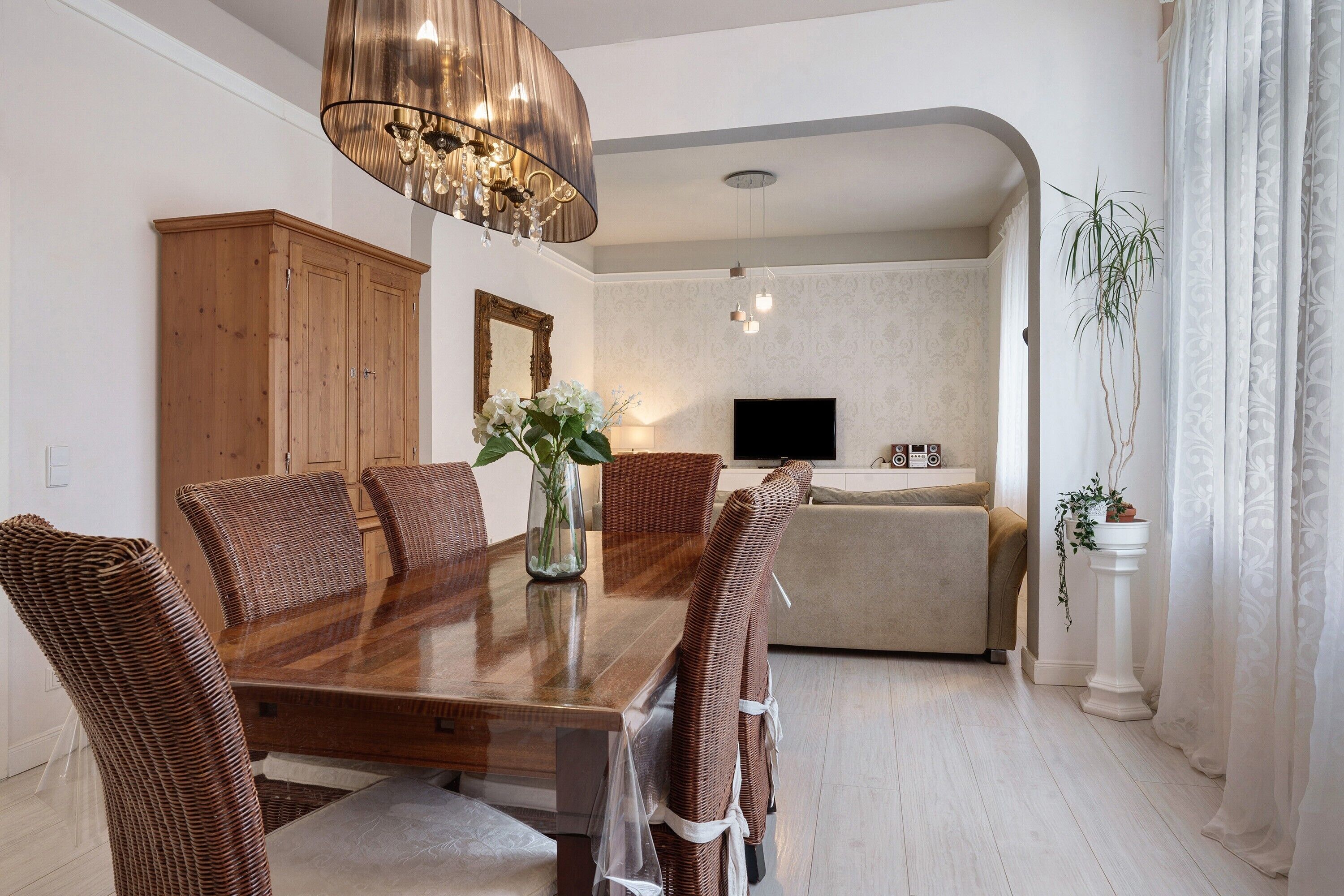 A dining area with a wooden table and wicker chairs, a chandelier, a television in the background, and a potted plant.
