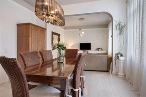 Uma sala de jantar com uma mesa de madeira e cadeiras de vime, um candelabro, uma televisão ao fundo e um vaso com planta.