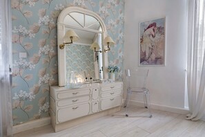 Casa de banho com papel de parede floral, toucador com espelho e uma cadeira transparente.