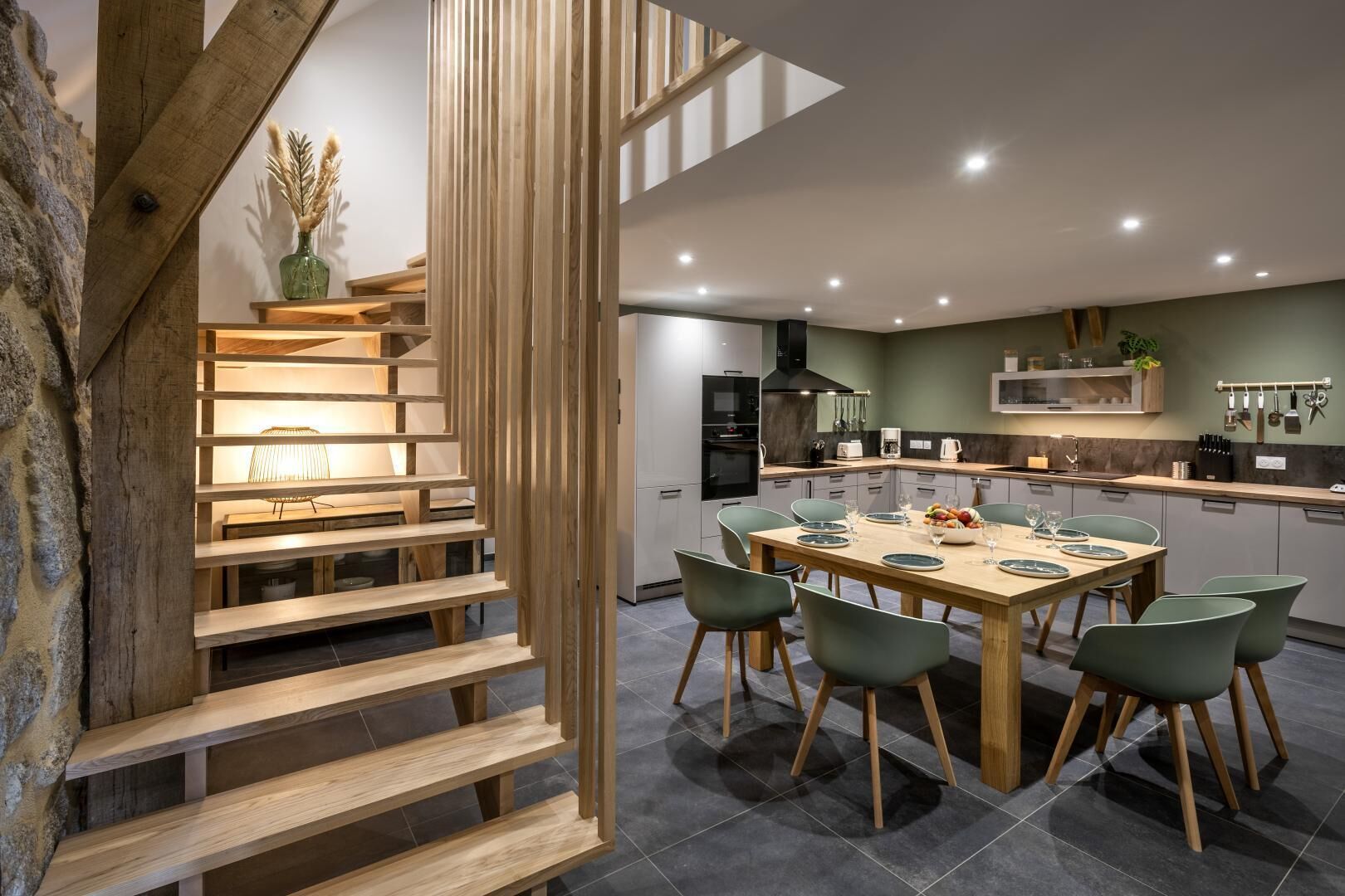 A modern kitchen with a dining area, featuring a wooden staircase, a dining table with chairs, and a kitchen island.