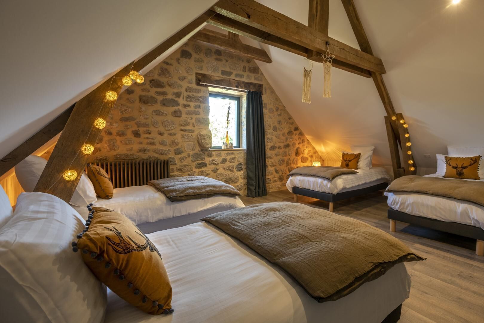 Een gezellig slaapkamer op zolder met stenen muren, houten balken en lichtjes. Er staan drie bedden met passend beddengoed en decoratieve kussens.
