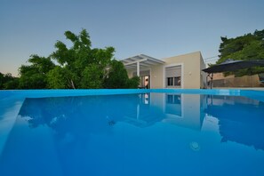 A swimming pool with a clear blue surface, surrounded by trees and a modern building with a covered patio. - Heli View Apartment with Sea View, Wi-Fi and Air Conditioning (Ermionida)