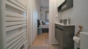 Un couloir étroit mène à une chambre où se trouvent un lit, une table de chevet et un petit tabouret. On aperçoit des toilettes sur la droite, et un lavabo est installé dans un coin.