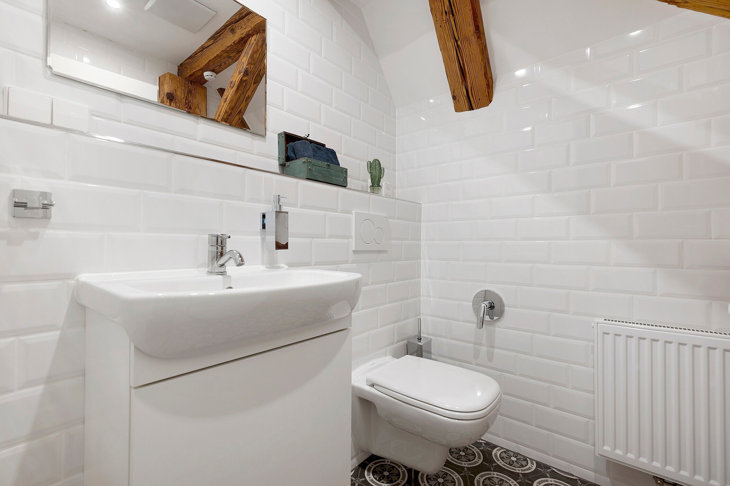 A bathroom with a white sink, toilet, and a mirror. There is a shelf with a towel and a cactus. The floor has a patterned design.