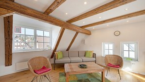 Una camera in stile loft con travi a vista in legno, un divano, due poltrone in vimini, un tavolino da caffè e ampie finestre che si affacciano sui palazzi circostanti.