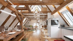 Une pièce aménagée dans les combles, avec un plafond orné de poutres en bois, une table à manger dressée avec des assiettes et des verres, un coin cuisine équipé d’un réfrigérateur et un escalier en colimaçon.