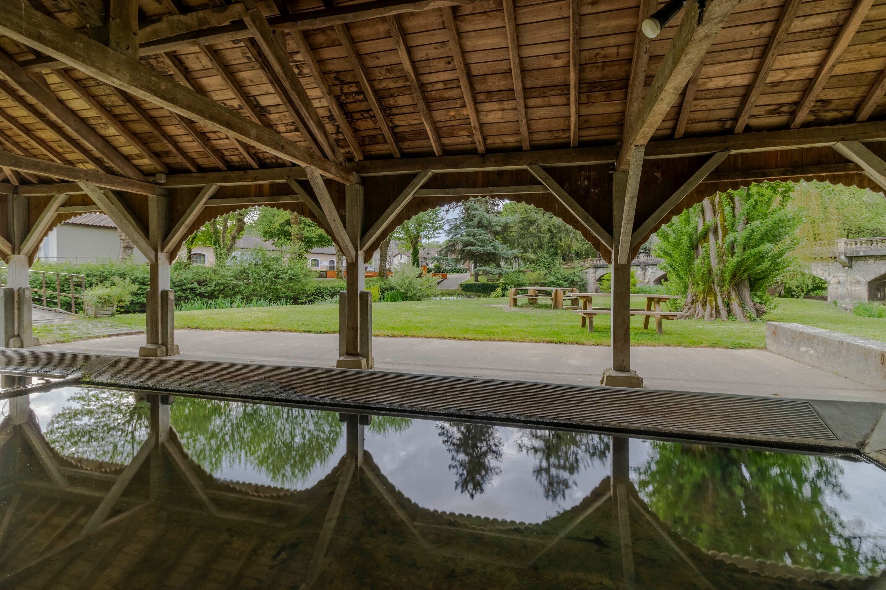A covered pavilion with a reflective floor, wooden beams, and a view of a park with benches and trees.