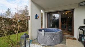 Ett utomhusbadkar står utanför ett hus, med ett område av grus, ett litet träd och en dörr som leder in.