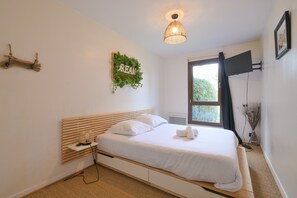 Ein Schlafzimmer mit Bett, Nachttisch, am Wand montiertem Fernseher, Fenster mit Vorhängen und einem Wanddekor.