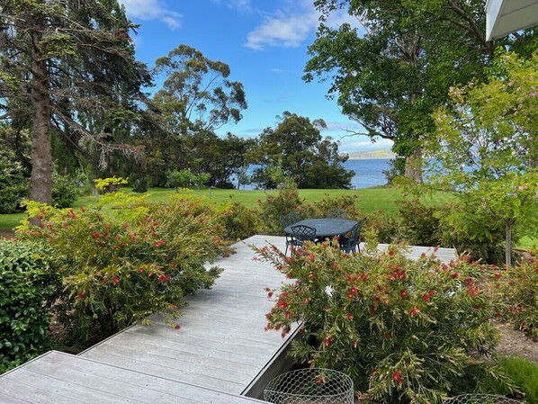 Una terraza de madera que conduce a una mesa y sillas junto a un lago, rodeada de abundante vegetación y árboles.