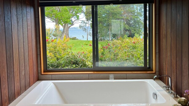 A bathroom with a large bathtub, wooden walls, and a window overlooking a garden.