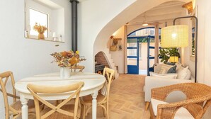 Zona pranzo con tavolo rotondo, sedie di legno e un vaso di fiori. Si nota una scala che conduce a un'altra stanza, un divano con cuscini e una lampada.
