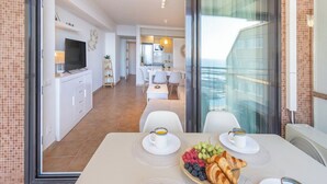 Ein modernes Hotelzimmer mit Balkon, einem Essbereich mit Frühstück und einer durchgehenden Tür erkennbaren Küche.