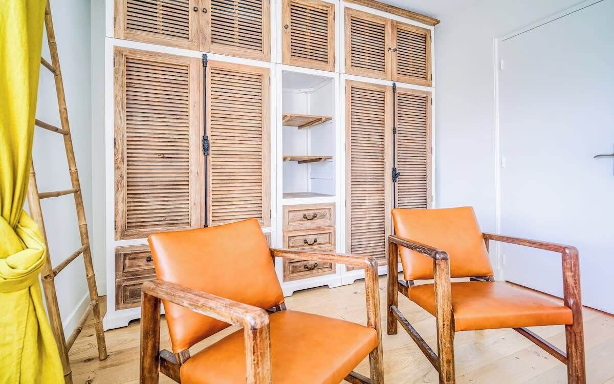 Een kamer met een grote houten garderobe, twee oranje stoelen en een geel gordijn.