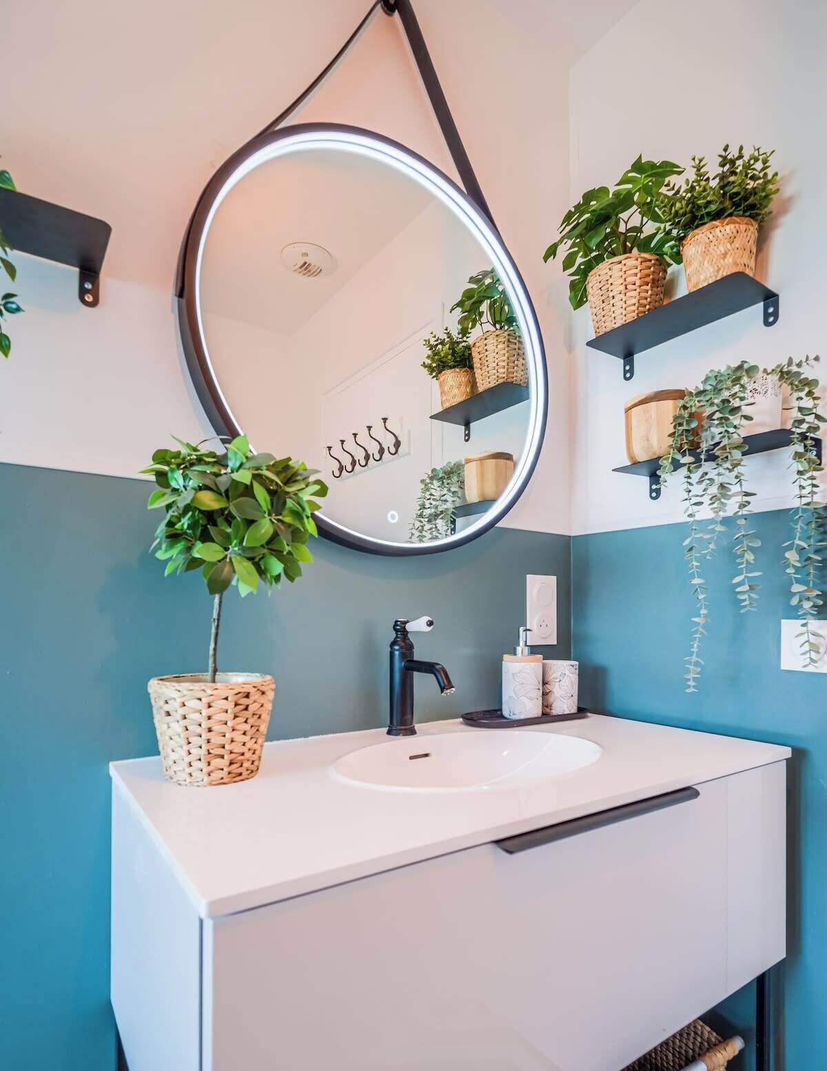 Een badkamer met een witte wastafel, een zwarte kraan en een spiegel met verlichting. Er staan planten op planken en aan de muur hangen haken.