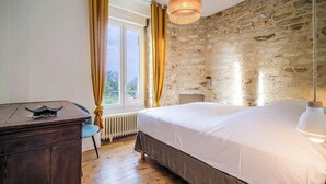 Un dormitorio con pared de piedra, un escritorio de madera, una cama con ropa de cama blanca y una ventana con cortinas.