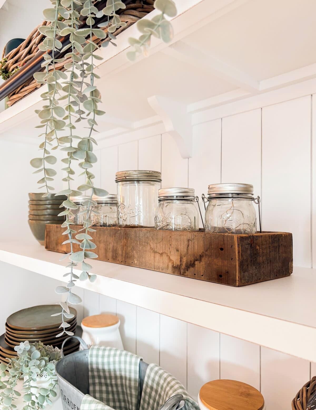 A kitchen shelf with glass jars, a wooden block, and hanging eucalyptus leaves.