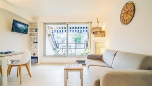A living room with a sofa, a TV mounted on the wall, a dining table with chairs, and a large window with a view of the outdoors. - Vacances à pied studio charmant à 1min des plages (Carnac)