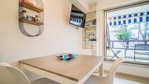 A room with a wooden table and white chairs, a TV mounted on the wall, and a window with a view of outdoor furniture. - Vacances à pied studio charmant à 1min des plages (Carnac)