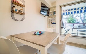Una habitación con una mesa de madera y sillas blancas, un televisor montado en la pared y una ventana que ofrece vistas a muebles de exterior.