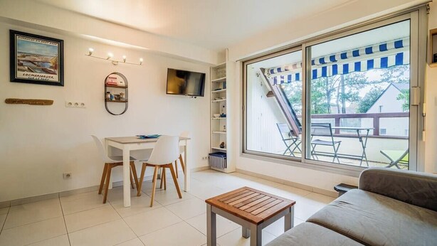 A modern living room with a sofa, a wooden coffee table, a dining table with chairs, a TV mounted on the wall, and a large window with a view of the outside. - Vacances à pied studio charmant à 1min des plages (Carnac)
