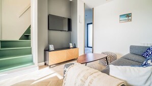 Un soggiorno moderno con un divano grigio, un tavolino in legno e una TV montata a parete. Si intravede una scala con gradini verdi a sinistra.