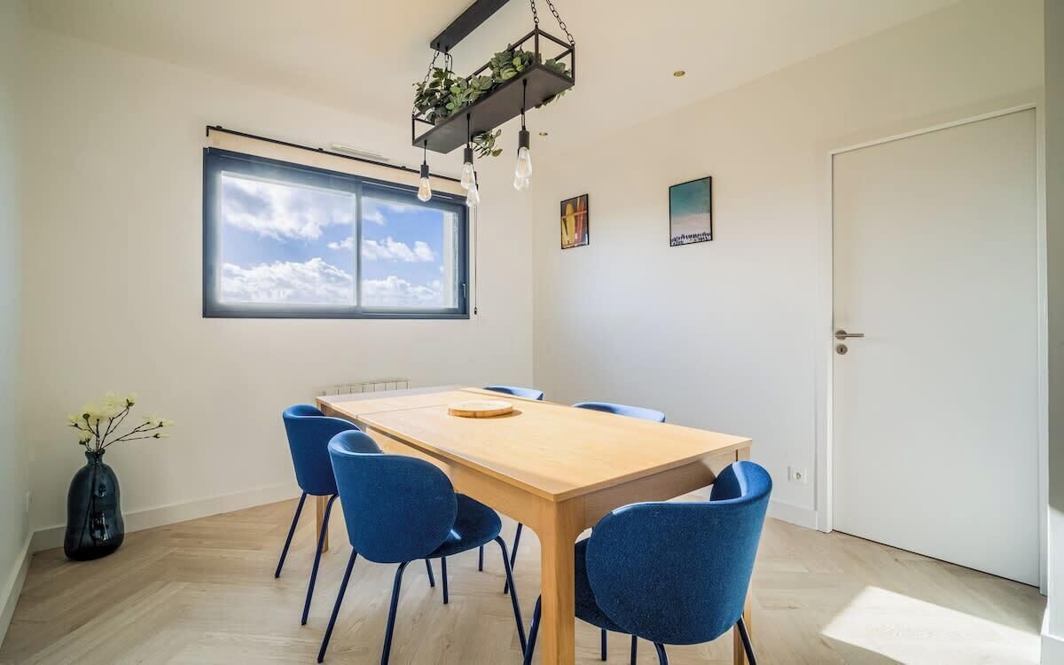 Un espace repas moderne avec une table en bois et des chaises bleues, une grande fenêtre donnant sur le ciel et une porte sur la droite.