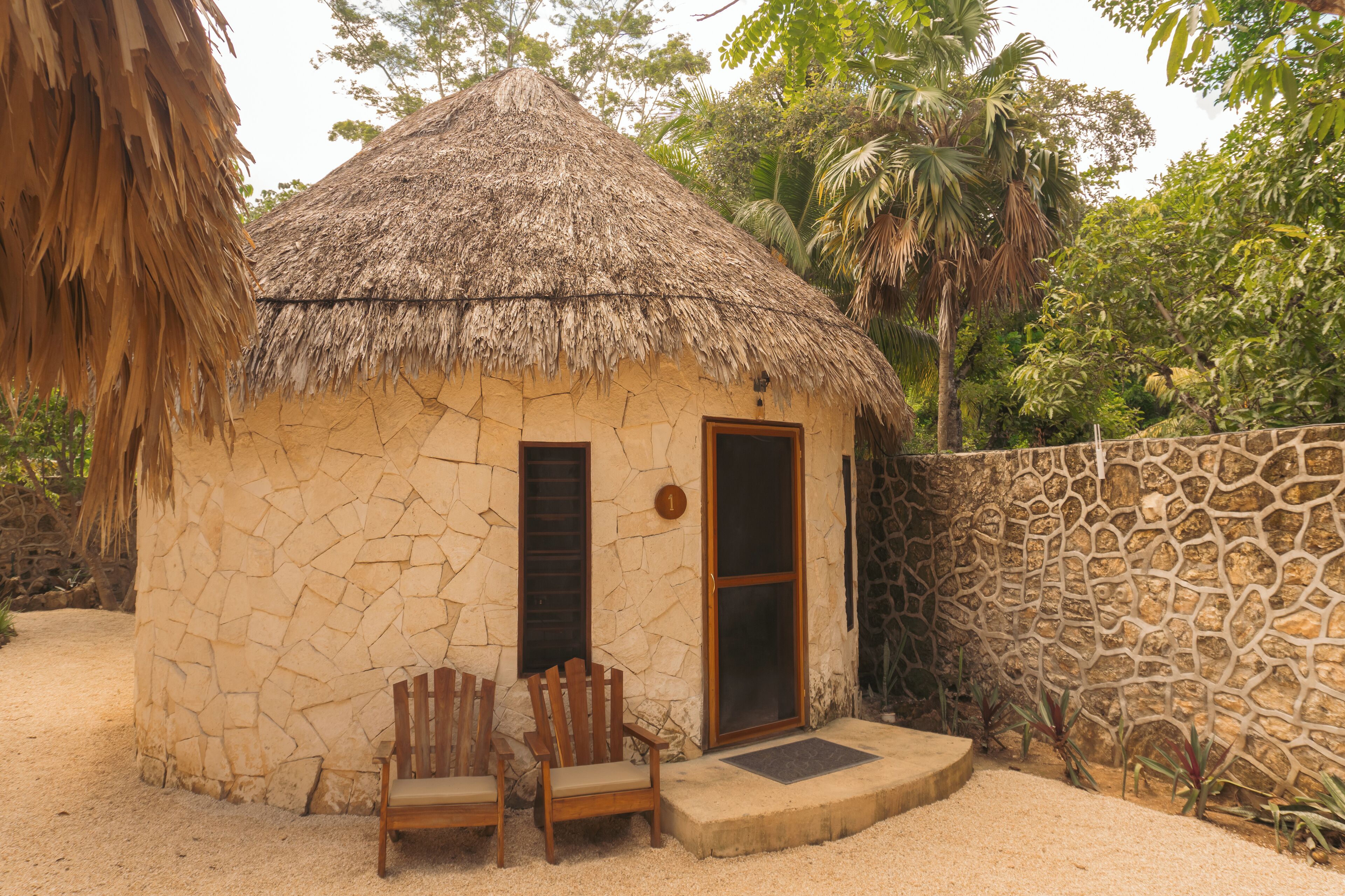 Une petite maison en pierre avec un toit de chaume, deux chaises en bois à l’extérieur et un mur de pierre.