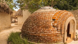 Una construcció amb cúpula de maó tradicional i teulat de palla, envoltada de vegetació i una paret de pedra.