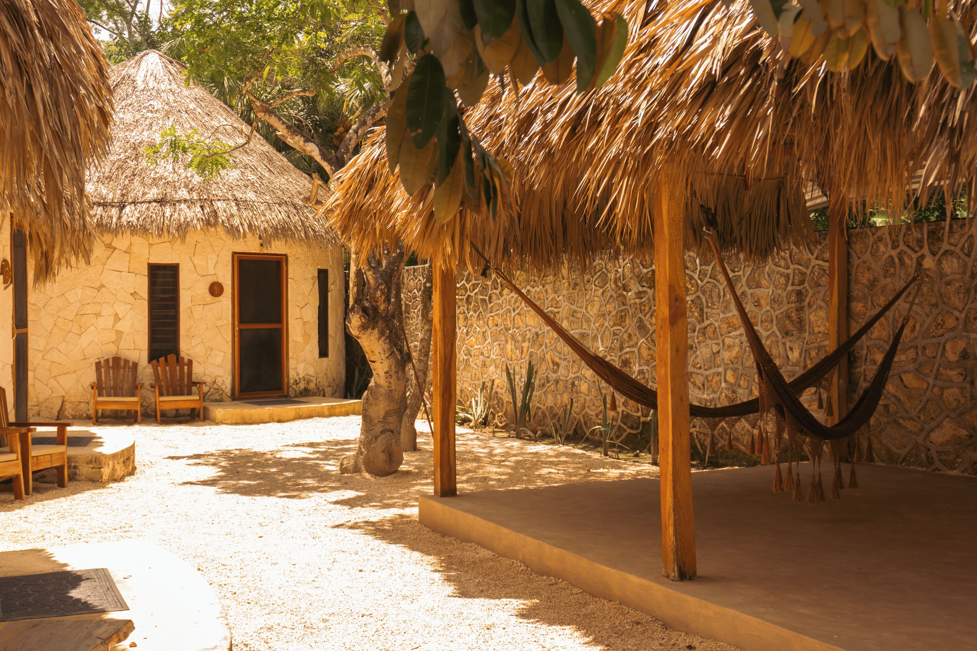 Une hutte au toit de chaume, avec un mur en pierre, des chaises en bois et un hamac.