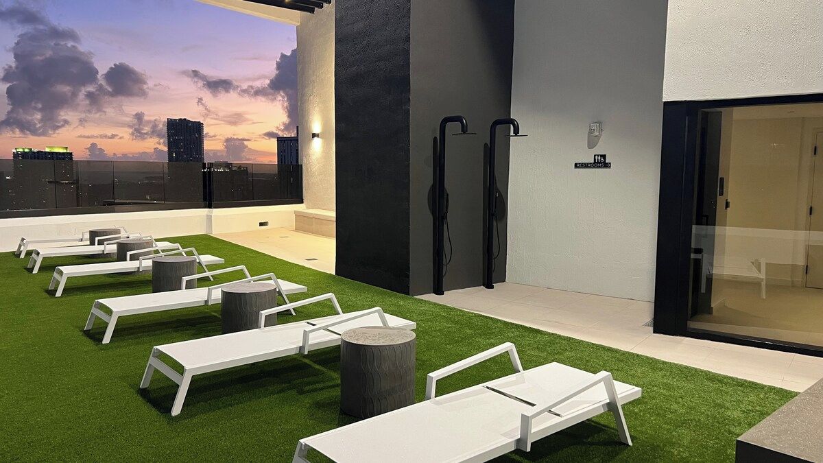 A rooftop lounge area with white lounge chairs, a grassy surface, and a cityscape view.