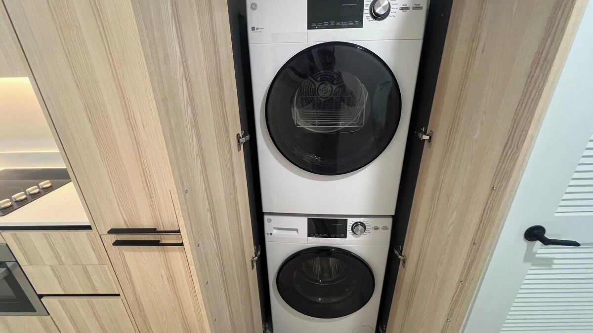 A compact washer and dryer unit in a modern kitchen with wooden cabinetry and a glass door.