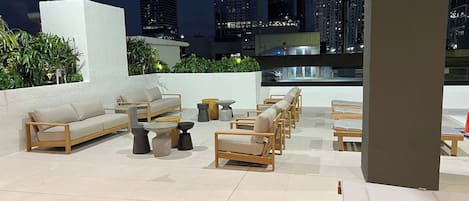 A rooftop terrace with outdoor seating, including sofas and chairs, and a view of city buildings at night.